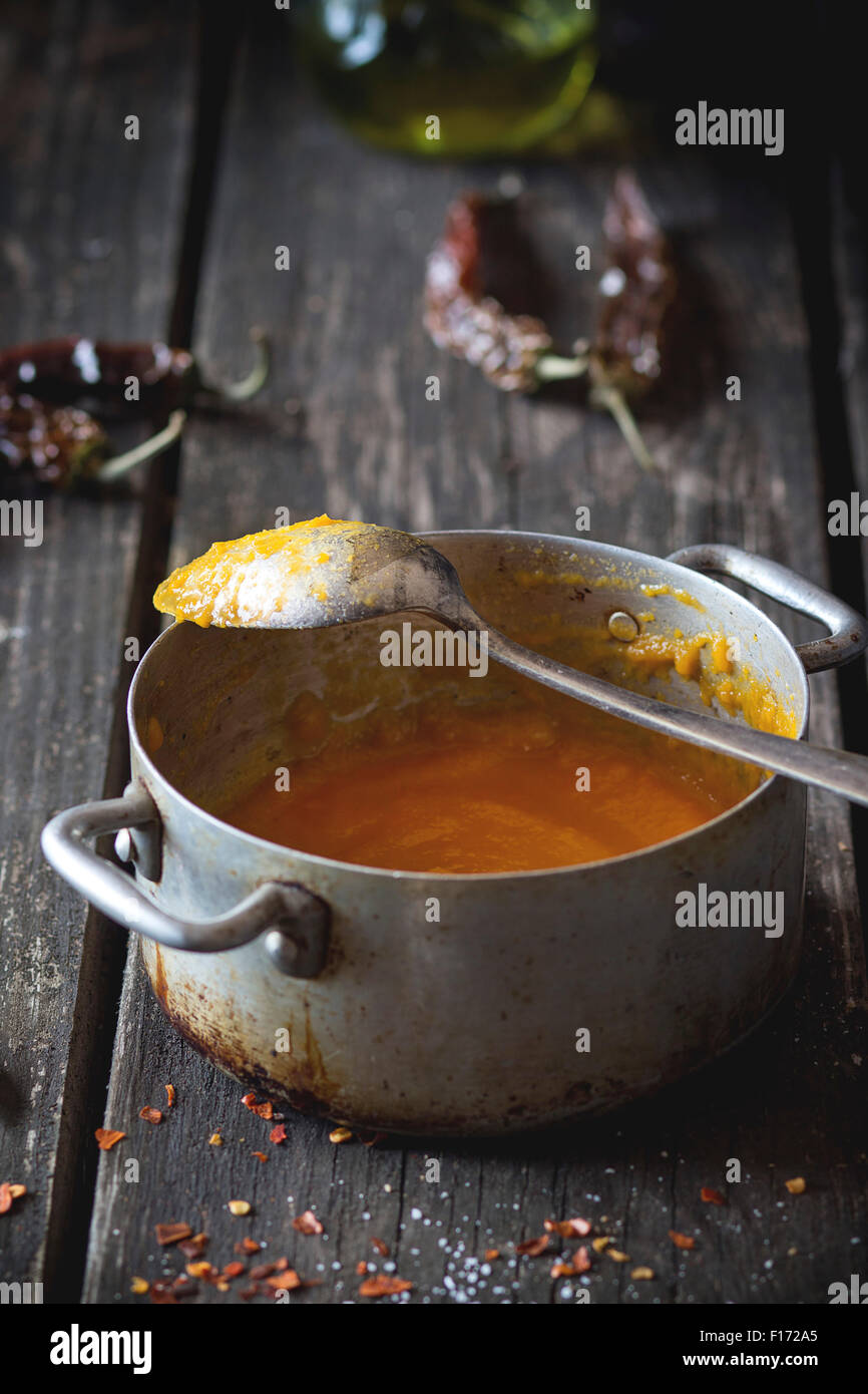Aluminum pan with remains of carrot cream soup with red hot chili pepper. Over old wooden table with vintage spoon and dry peppe Stock Photo
