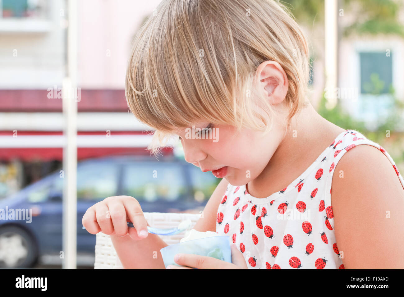 Blond Caucasian baby girl eats frozen yogurt, close up outdoor portrait with natural light Stock Photo