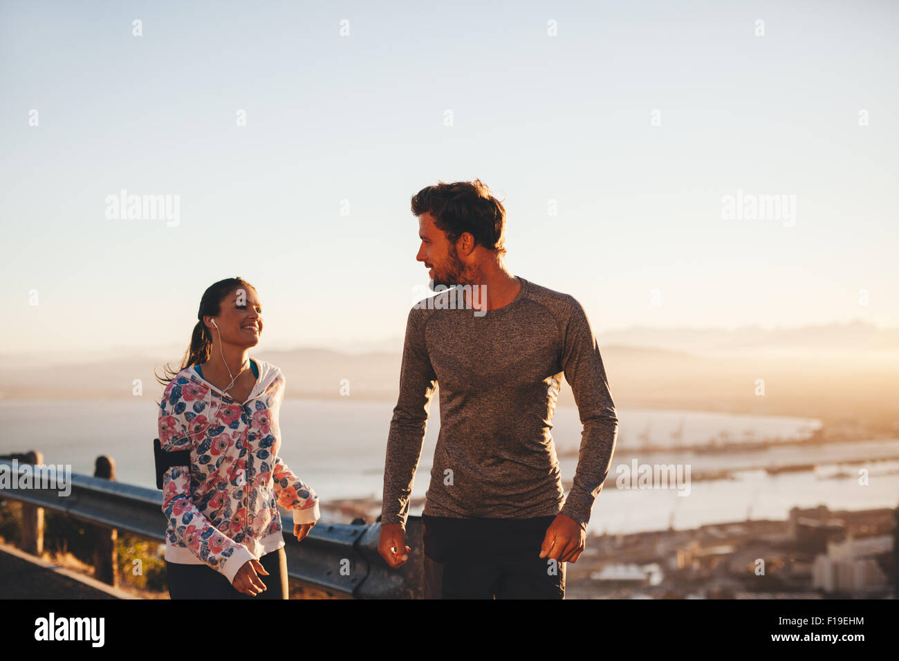 Image of fit young man and woman jogging on country road, looking at each other. Runners enjoying morning run. Stock Photo