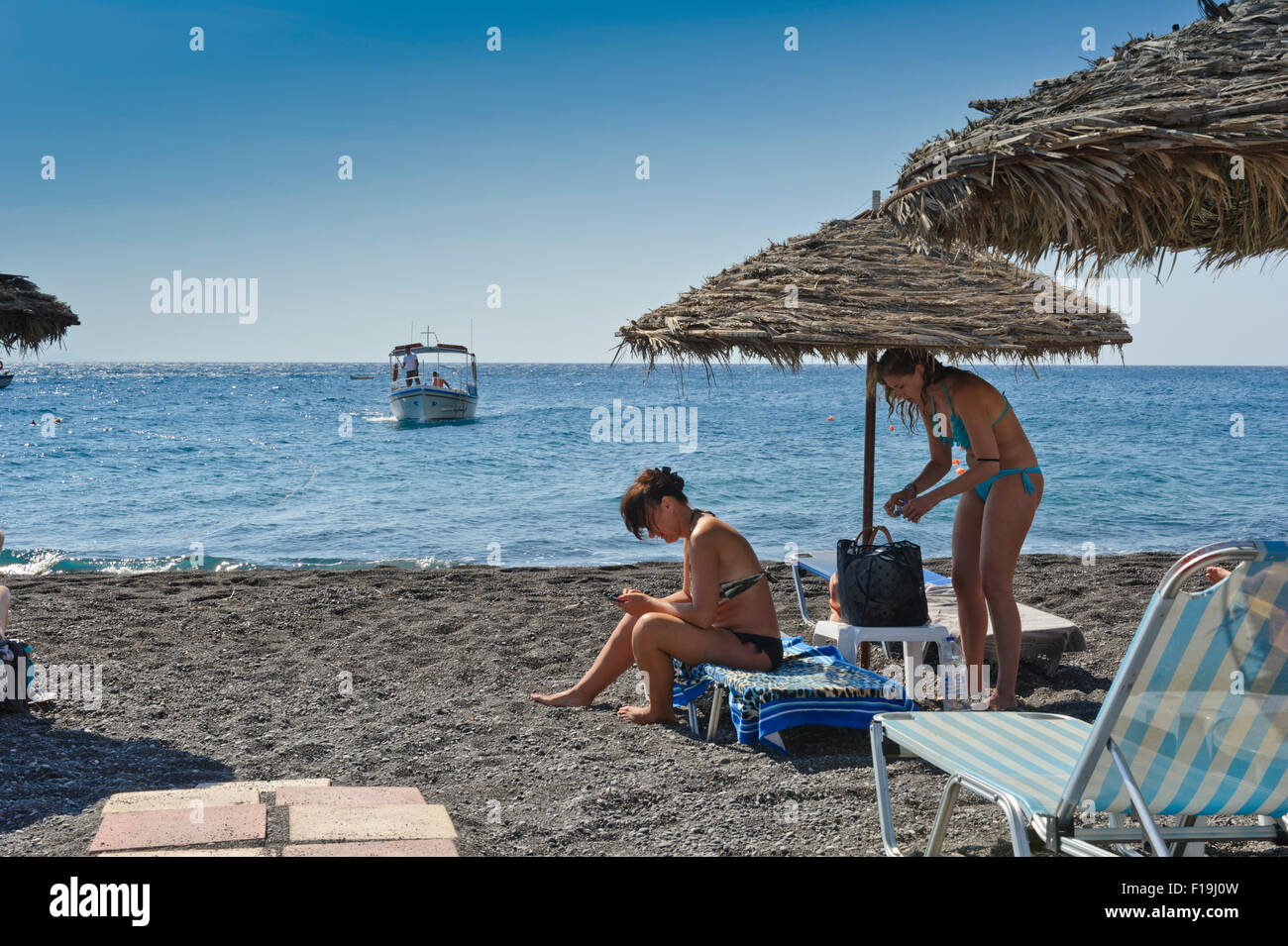 Two female sunbathers under a straw umbrella while a small boat approaching the beach in Kamari, Santorini, Greece. Stock Photo