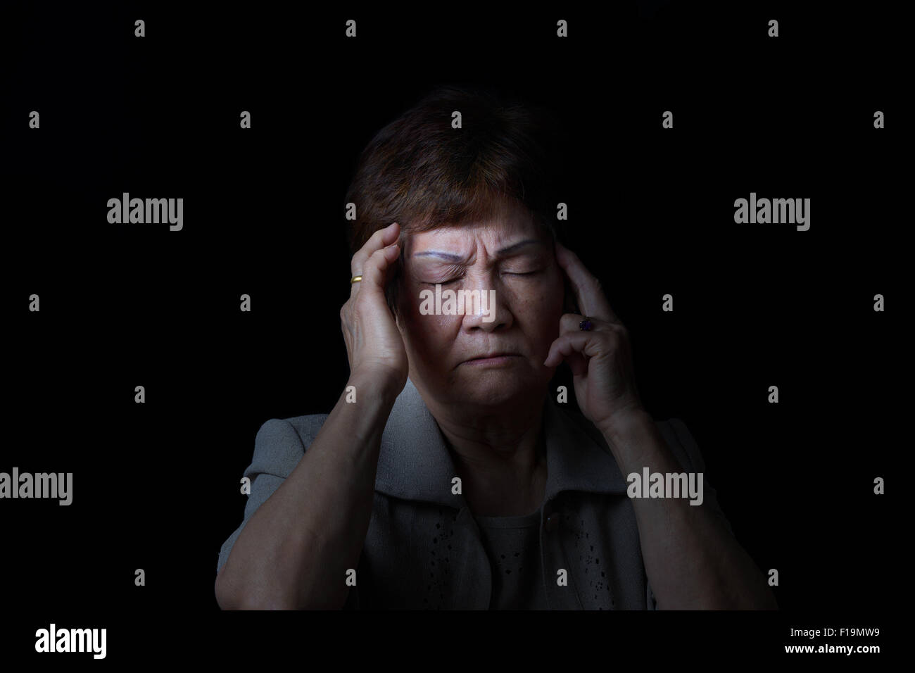 Senior woman touching the sides of her head with fingers while displaying pain on black background. Stock Photo