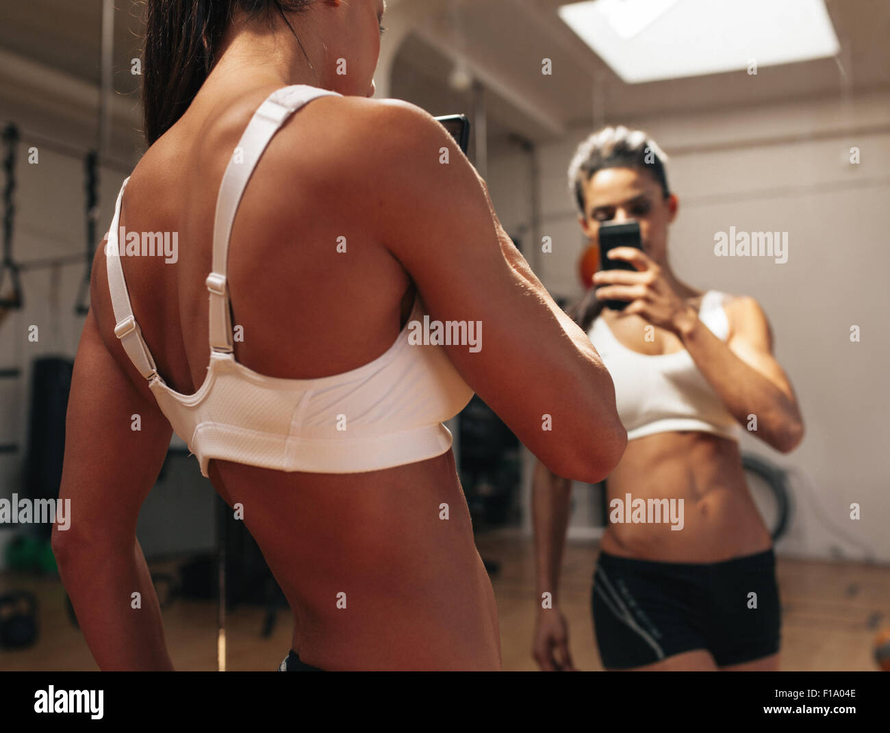 Sporty young woman taking a picture of herself in a mirror. Fitness model taking a selfie in front of a mirror in gym. Stock Photo