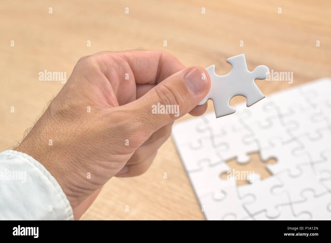 Businessman hand putting a missing piece and solving blank white jigsaw puzzle placed on top of old wooden oak table, close up, Stock Photo