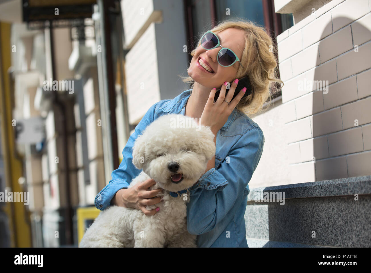 Cute blonde in sunglasses and a bright blue denim shirt emotionally talking on a cell phone. Bichon Frise white dog sitting on t Stock Photo