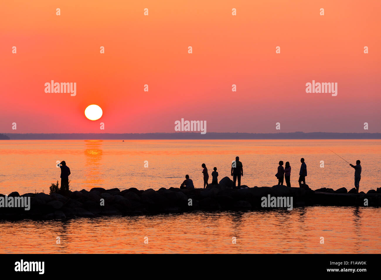 People silhouetted against a vibrant sky as the sun sets over Lake Simcoe. Willow Beach, Ontario, Canada. Stock Photo