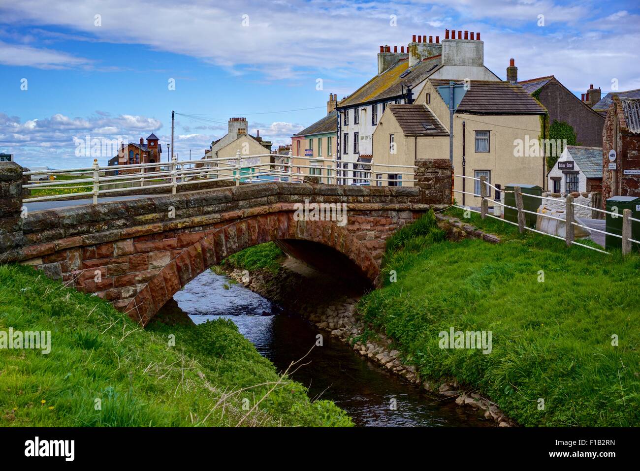 A red stone bridge across the River Ellen in Alonby, Cumbria, England., with assorted painted houses, blue sky, green grass. Stock Photo