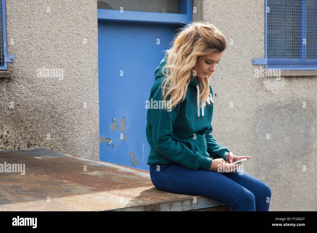 Teenage girl sitting on a wall outside wearing a green sweatshirt. Stock Photo