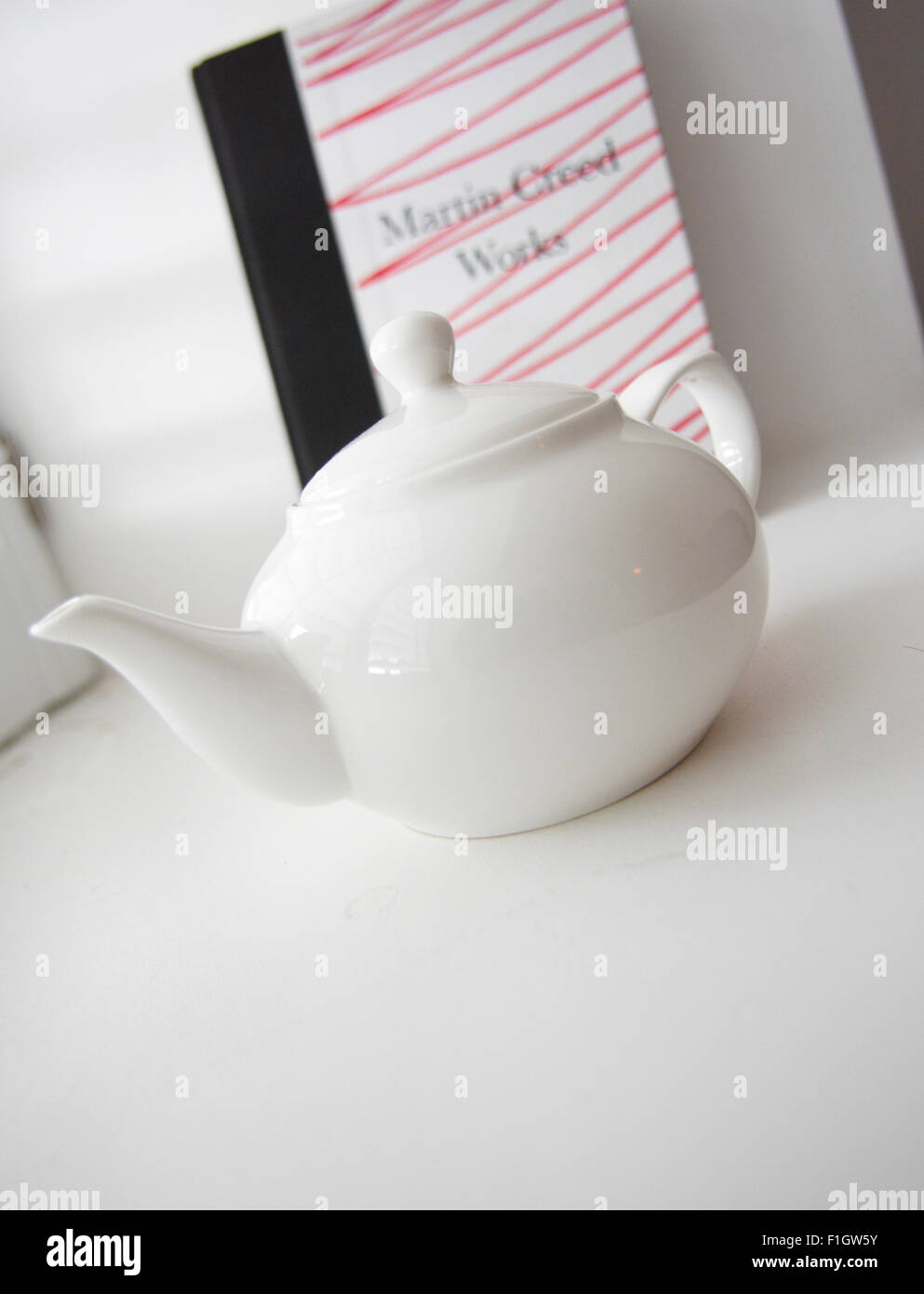 A small white teapot in a plain white location, in front of a book on