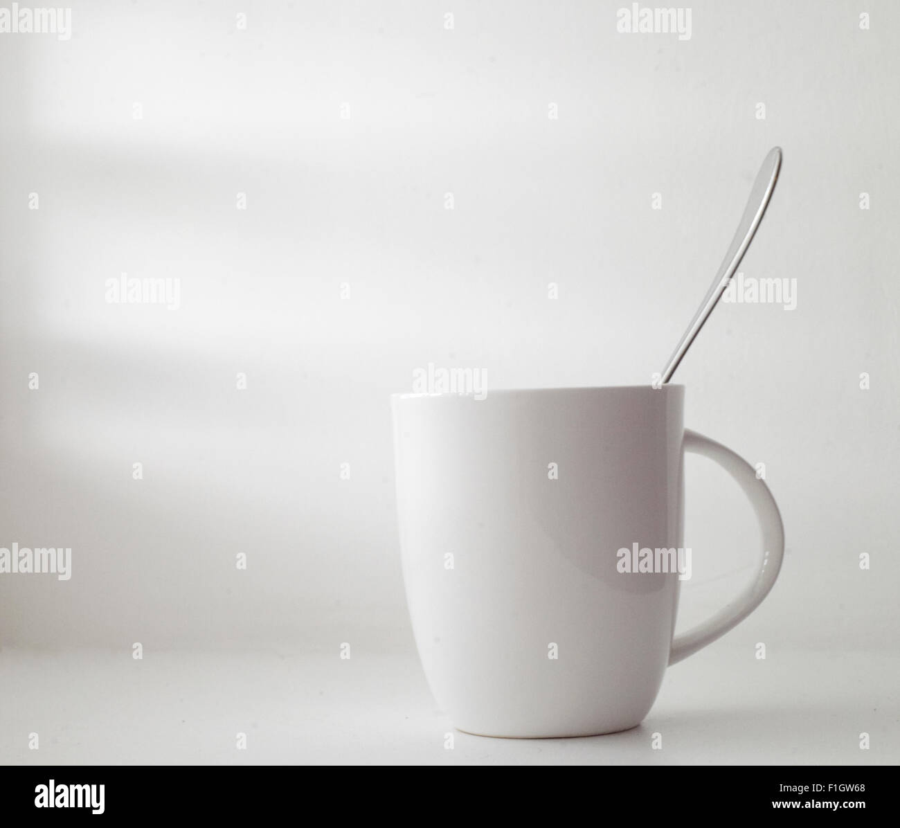 A white drinks mug with a silver spoon in it. Taken against a white wall. Stock Photo