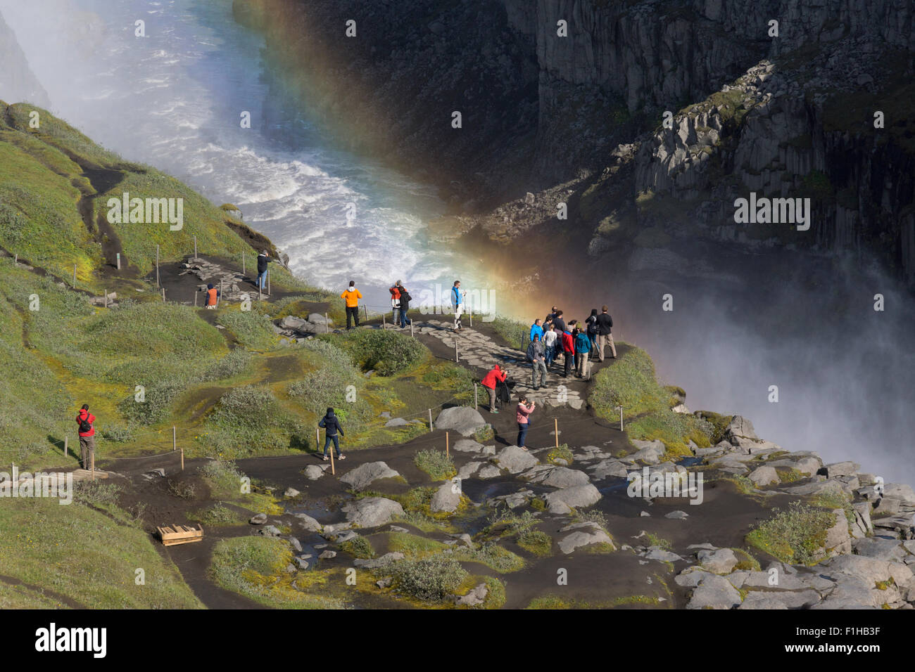 Tourists walking net to and viewing the Dettifoss waterfall with a rainbow behind in the mist, Vatnajökull National Park, northeast Iceland Stock Photo