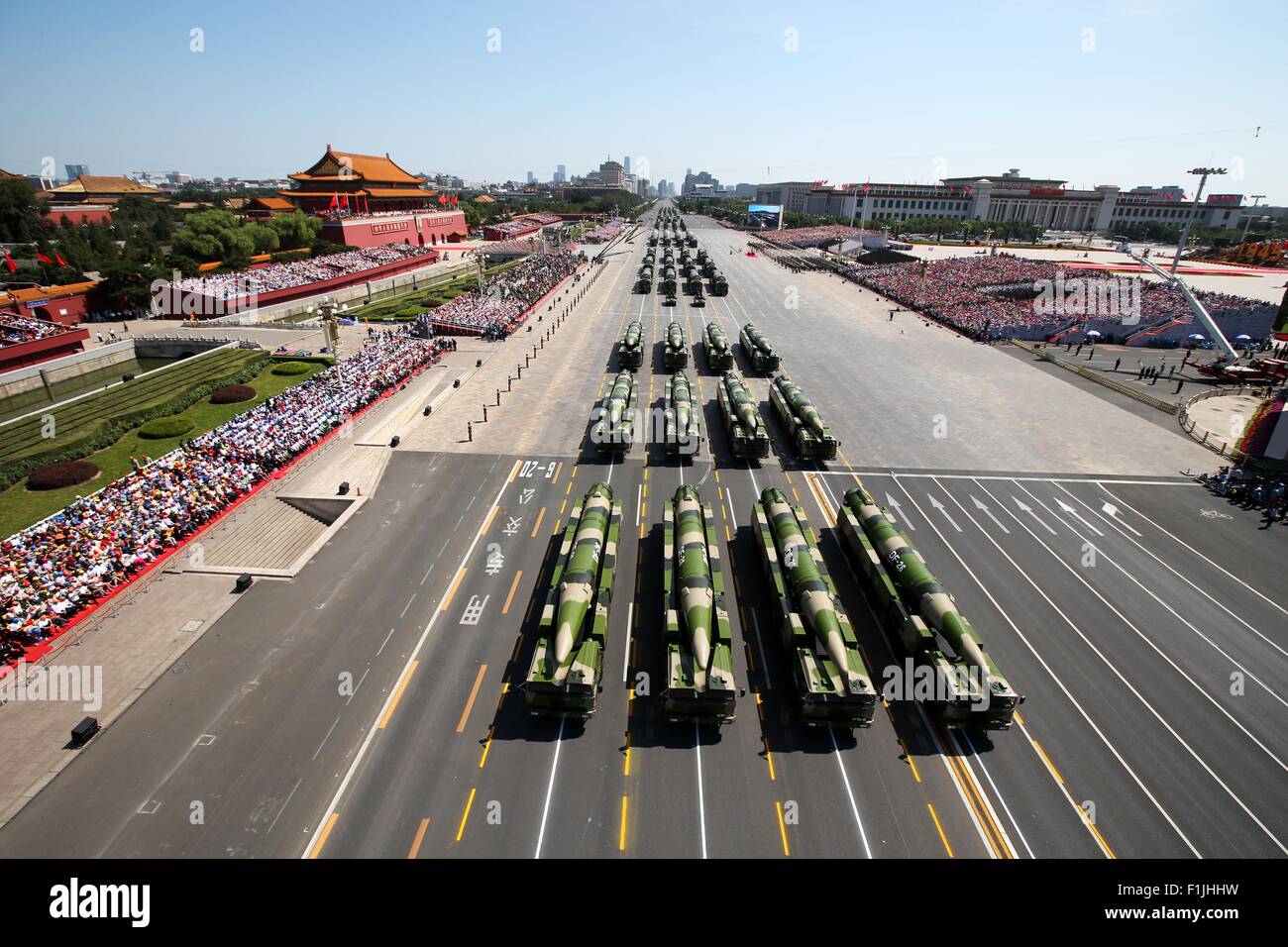 Military vehicles, including missile launchers, are parade ground, with a large crowd and spectators along the sides, and historic Chinese architecture and government buildings in the background.