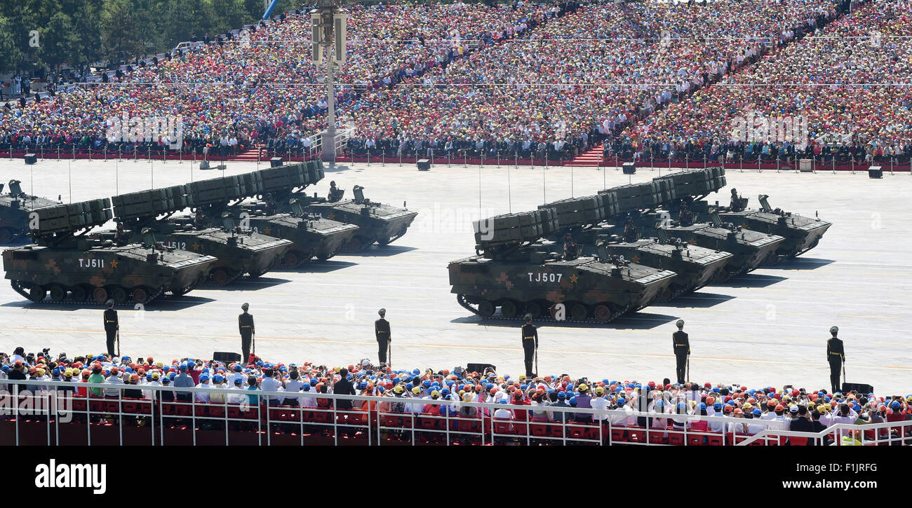 Military vehicles with missile launchers are parked in formation in front of a large seated crowd during a parade, with soldiers standing at attention in front of them.