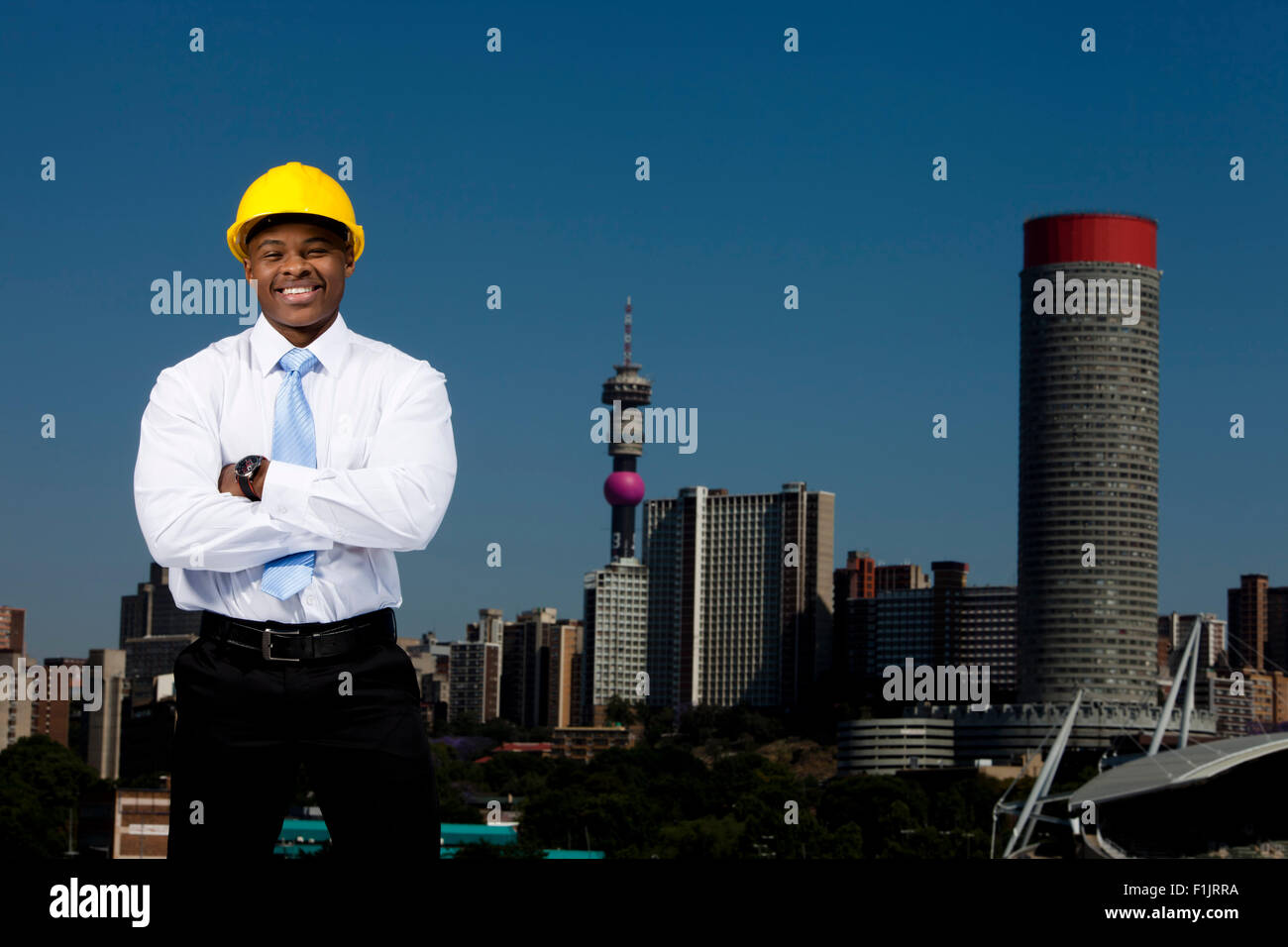 Smartly dressed business man wearing hardhat stands with arms crossed with cityscape in background Stock Photo