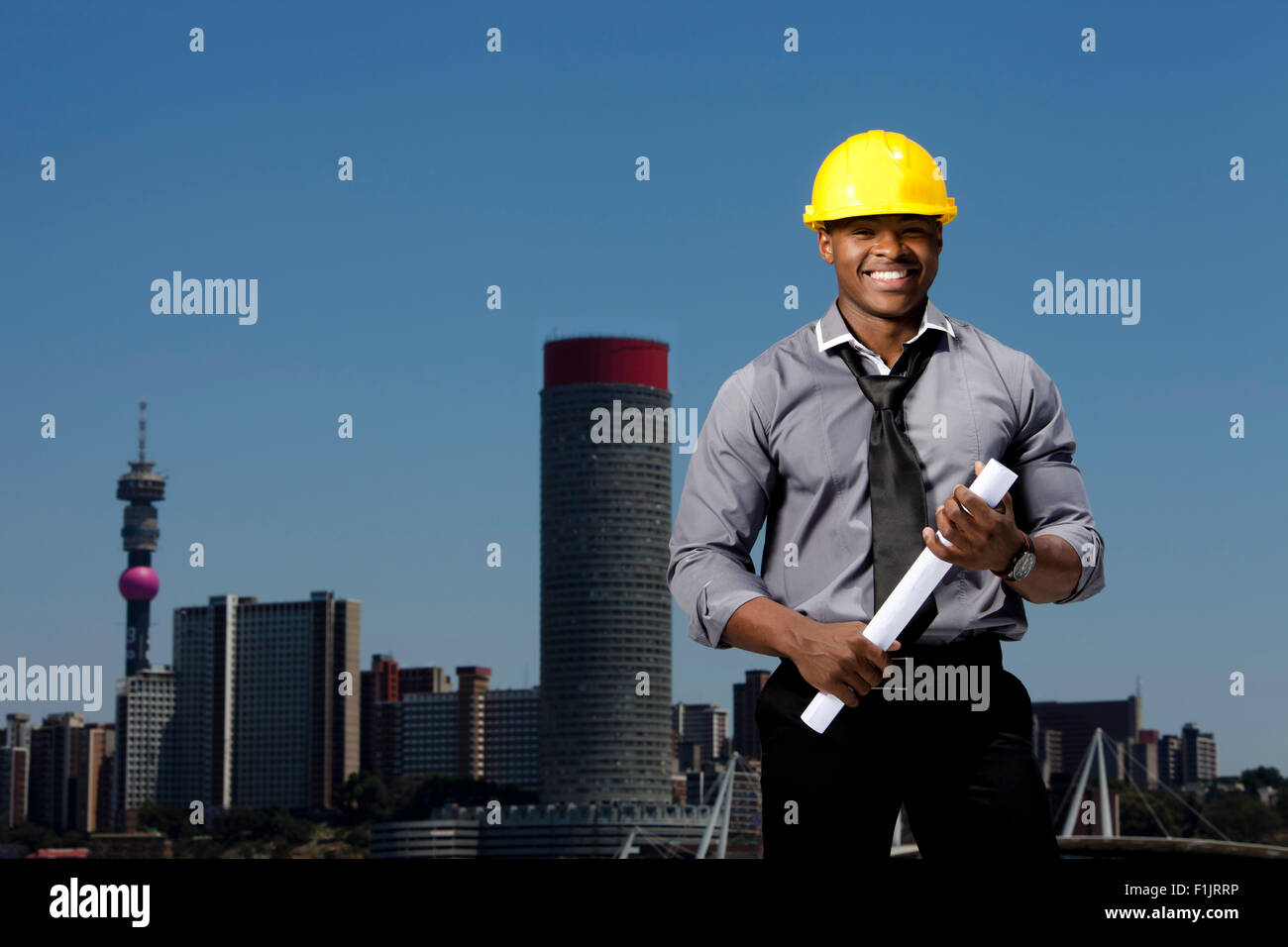 Smartly dressed business man wearing hardhat holds building plans while standing with arms crossed with cityscape in background Stock Photo