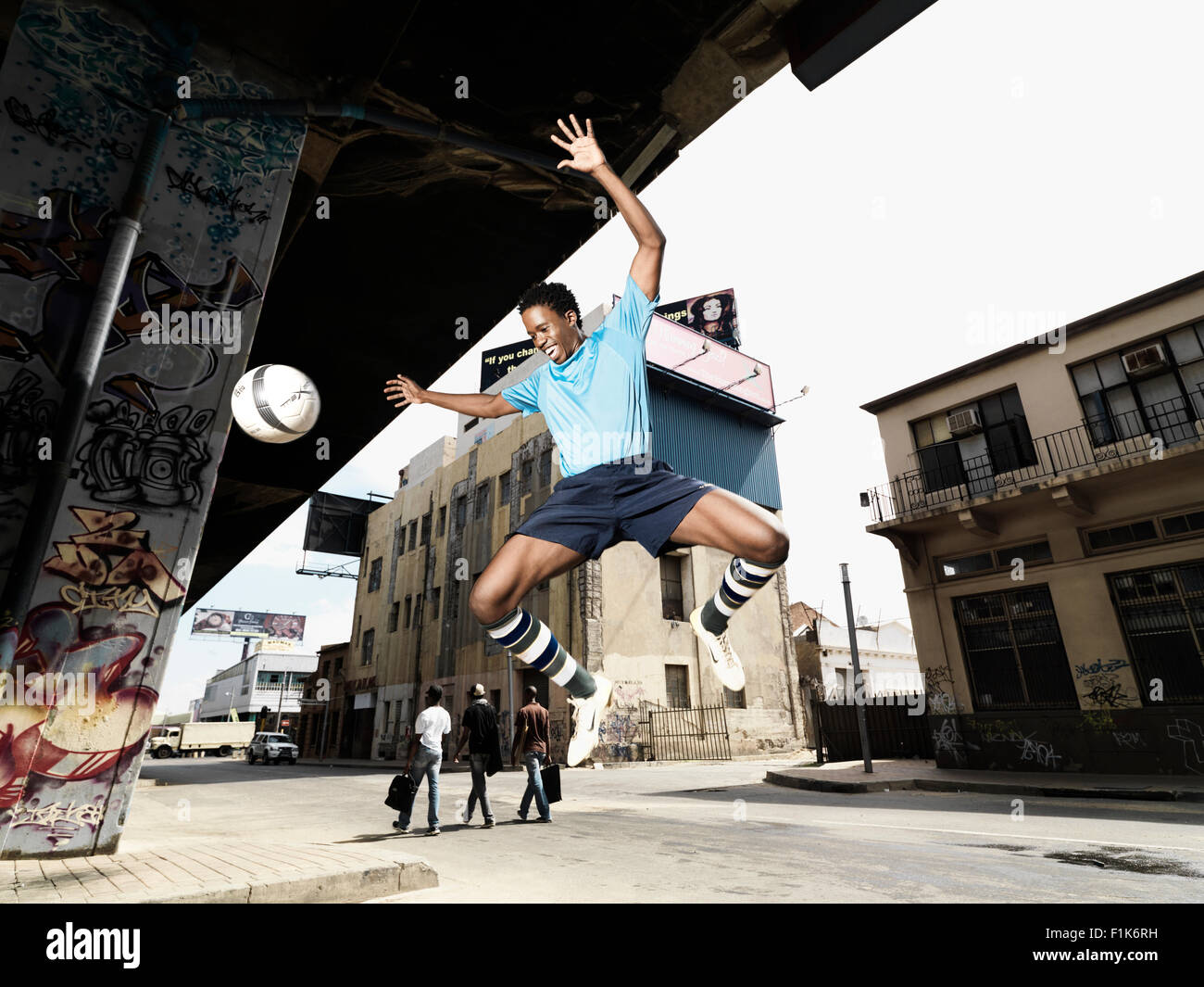 Black man jumping with soccer ball, city scene with people walking in the background Stock Photo