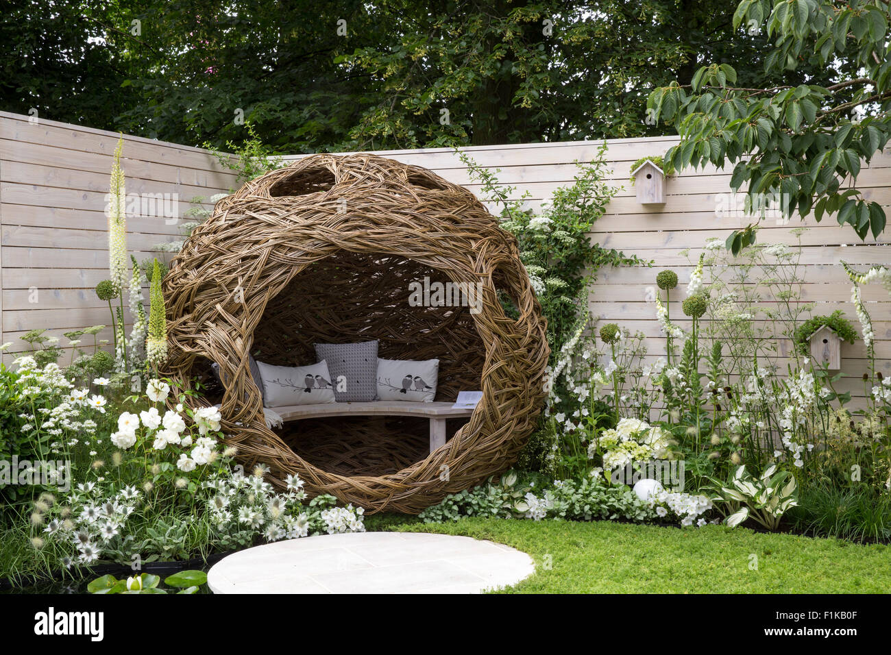 A small urban wildlife bird friendly garden chamomile lawn stepping stones and a woven willow pod with bench and cushions - white flowers Summer UK Stock Photo
