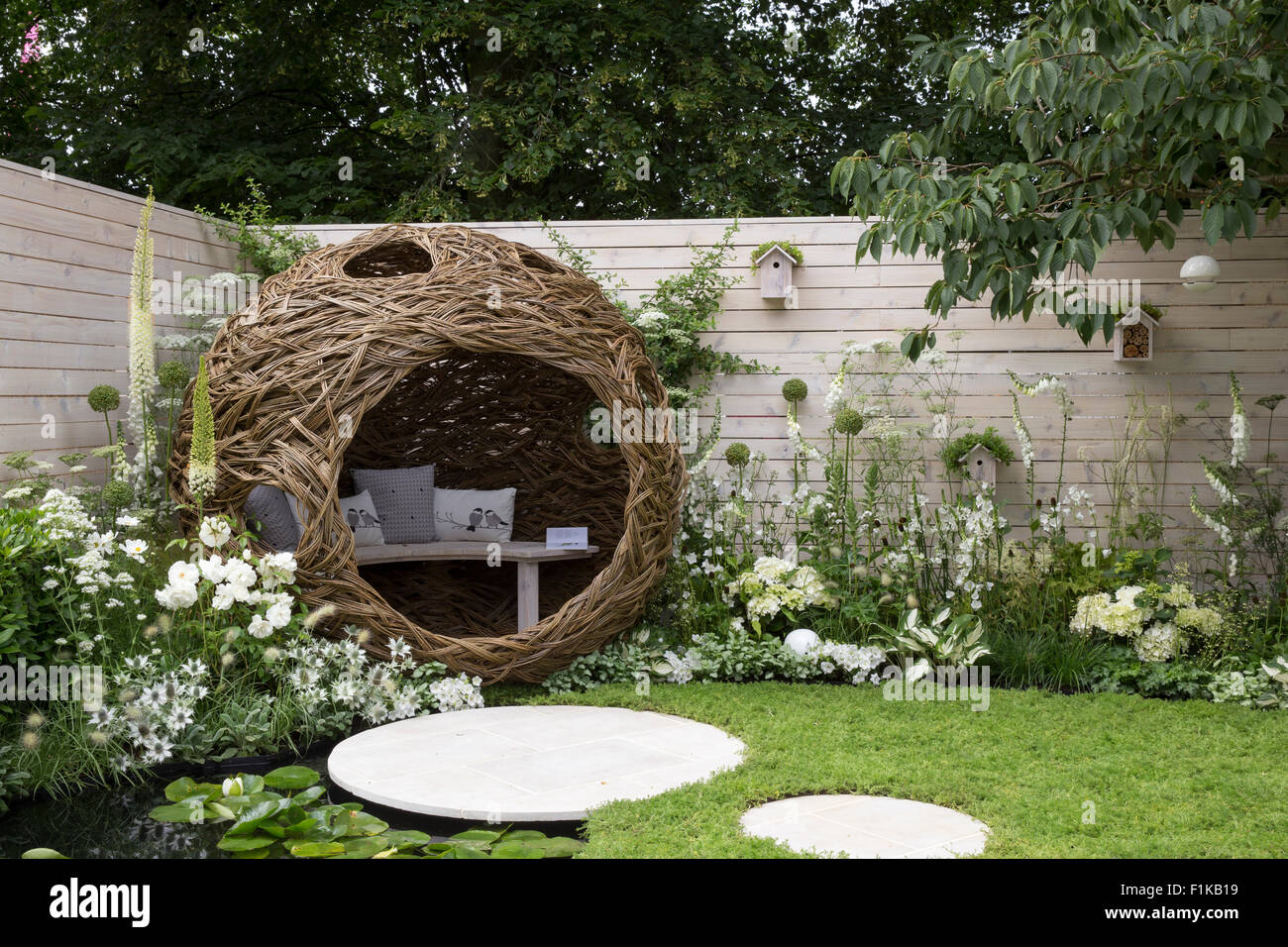 A small urban wildlife bird friendly garden chamomile lawn stepping stones and a woven willow pod with bench and cushions - white flowers Summer UK Stock Photo