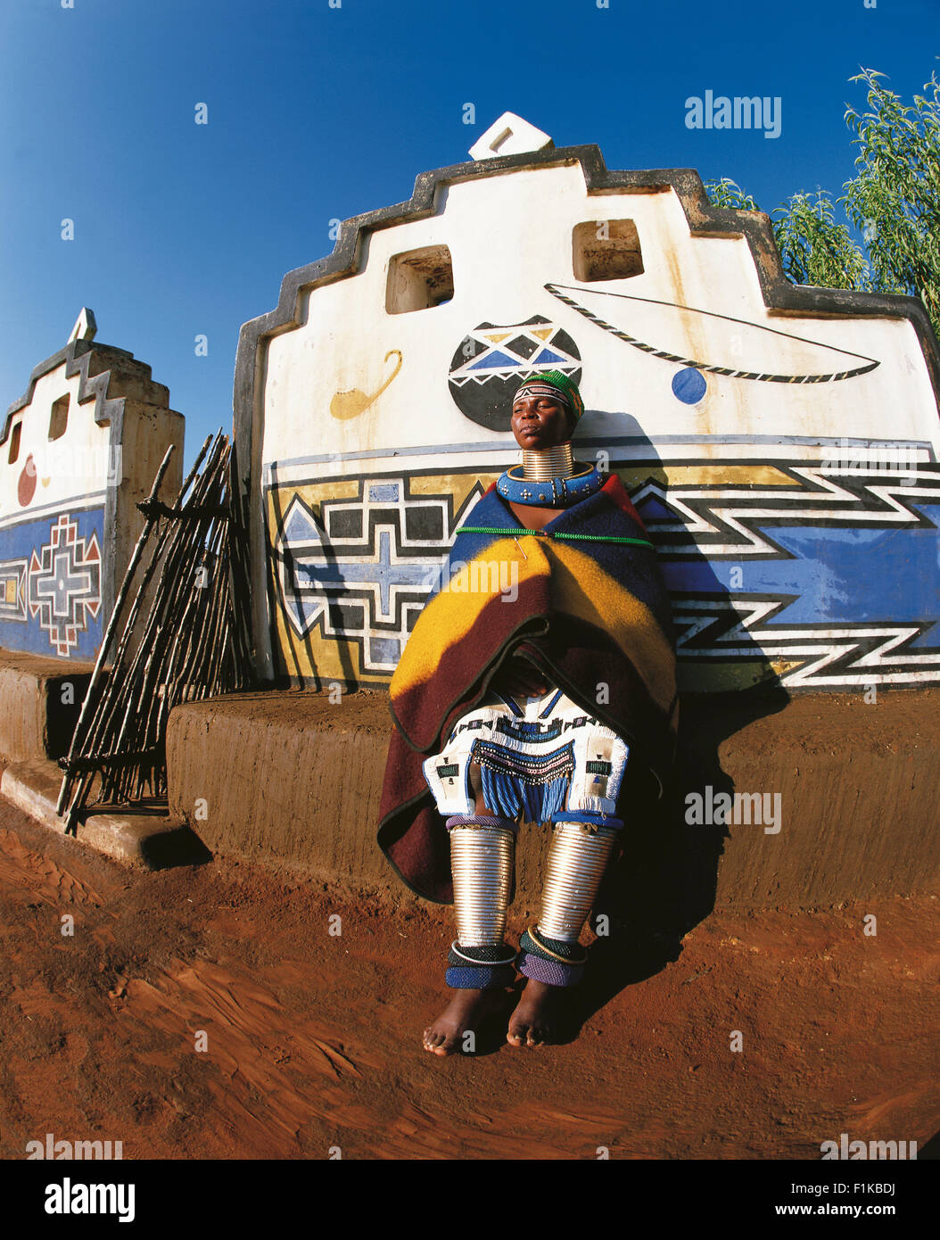 Ndebele woman in traditional dress leaning against wall of brightly coloured village. Botshabelo Mpumalanga South Africa Africa Stock Photo