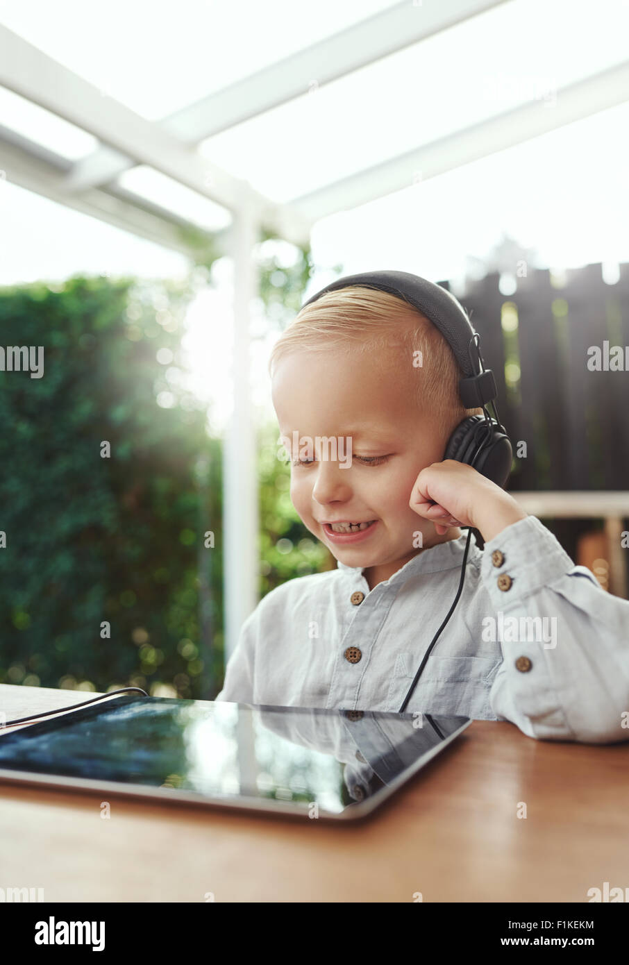 Little boy smiling in delight as he listens to music downloaded on his tablet-pc using stereo headphones while sitting outdoors Stock Photo