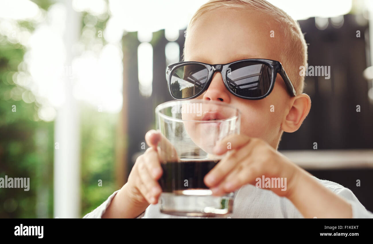 Cute hipster little boy in over sized sunglasses belonging to his mother or father sitting sipping a beverage in a glass on an o Stock Photo