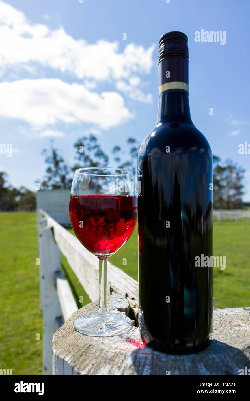 A red wine bottle and glass resting on a white fence in the country. Stock Photo