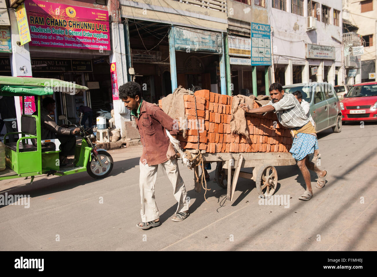 Delhi, India. Three men pushing a dilapidated wooden hand-cart with metal wheeels laden with new red bricks along a city road. Stock Photo