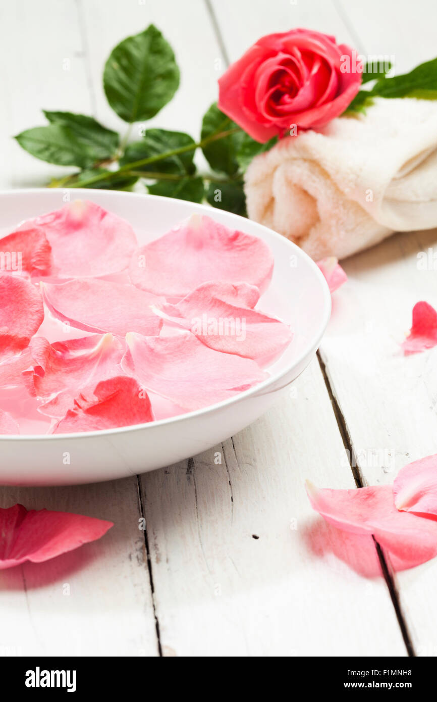 Spa concept with pink rose, rose petals in bowl of water and rolled towel on white wooden