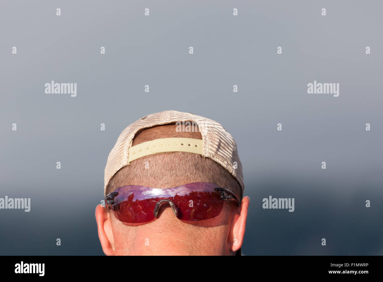 A man wears his sunglasses on the back of his shaved head with a hat that gives the illusion of a face. Stock Photo