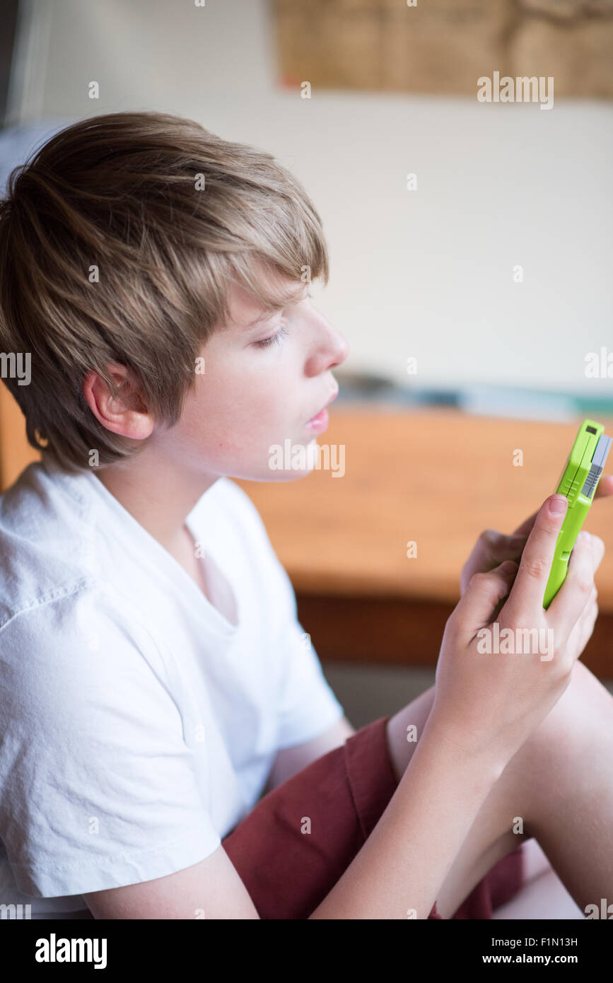White, blond-haired teenage boy in a white T shirt and shorts plays on a vintage  lime green Gameboy machine Stock Photo