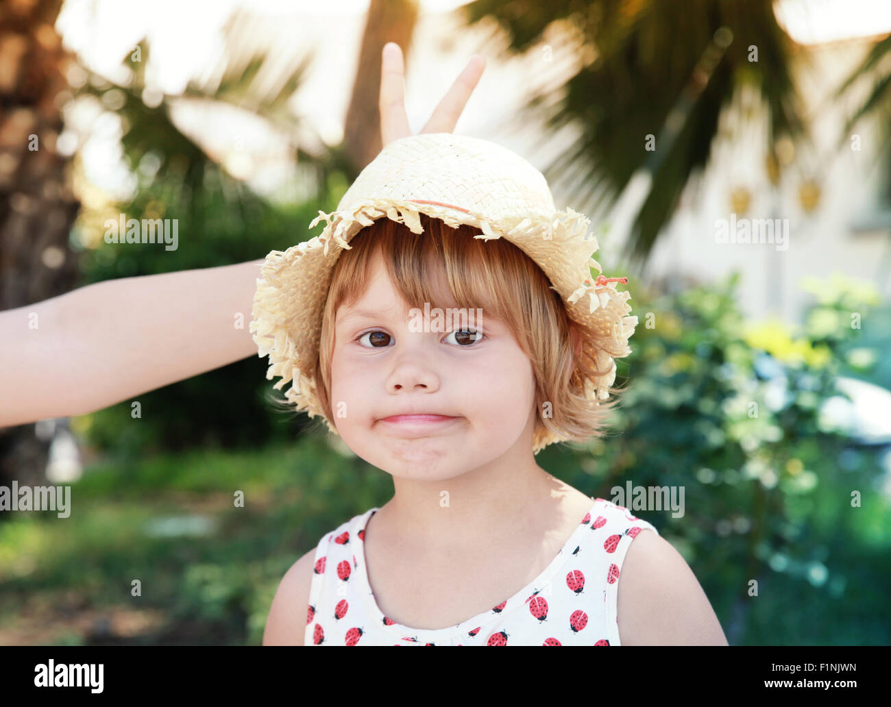 Cute Caucasian little girl in straw hat, other person makes horns with