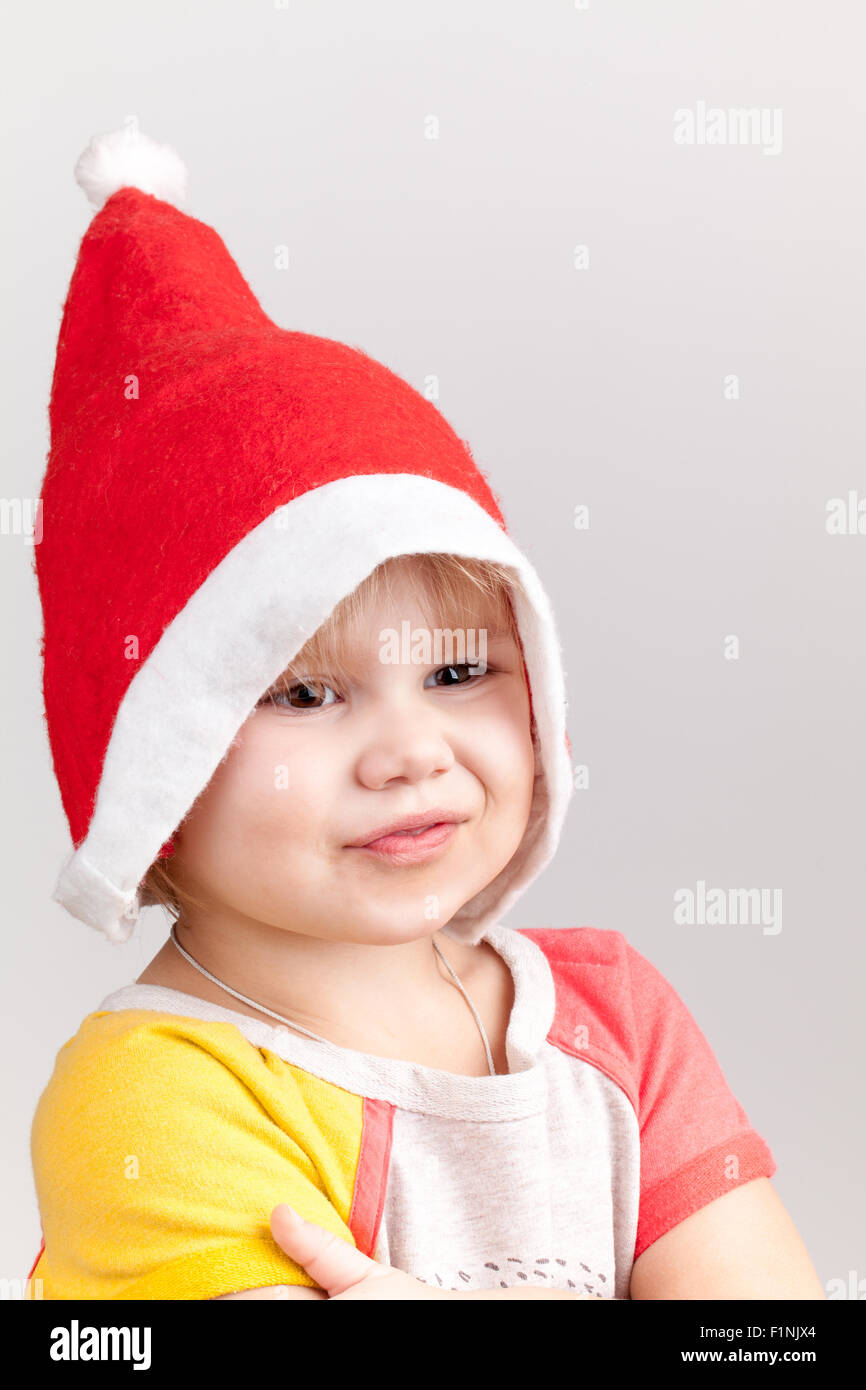 Cute smiling Caucasian little girl in red Santa hat on gray background, closeup studio portrait Stock Photo
