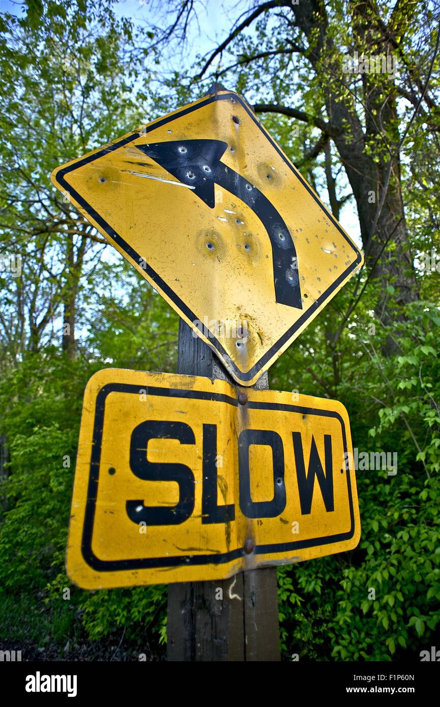 Bullet Holes Slow Down Sign Somewhere in the Wood. Damaged by Bullets Yellow Traffic Sign. Transportation Photo Collection Stock Photo