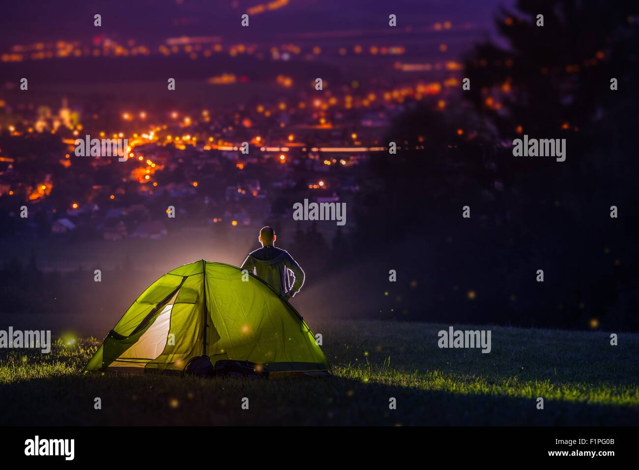 Countryside Camping with Scenic City View Down the Valley. Illuminated Cityscape at Night and the Camper with Illuminated Tent. Stock Photo