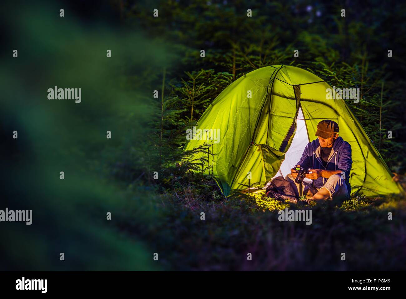 Overnight Forest Camping. Middle Age Caucasian Hiker with His Digital Camera and the Illuminated at Night Green Tent. Nighttime Stock Photo