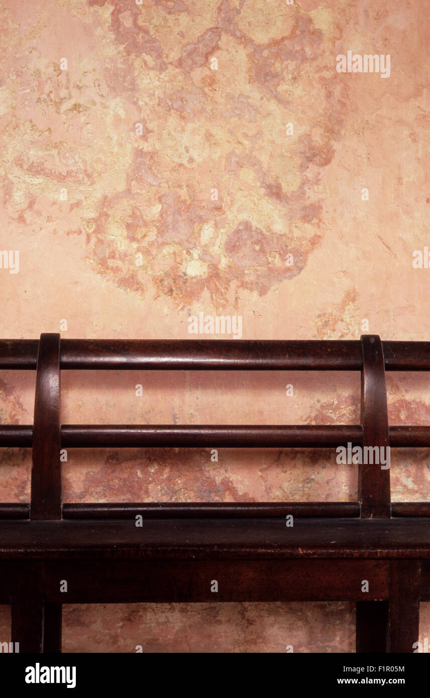 Dark mahogany wooden bench against faded pink wall with patched and peeling plaster Stock Photo