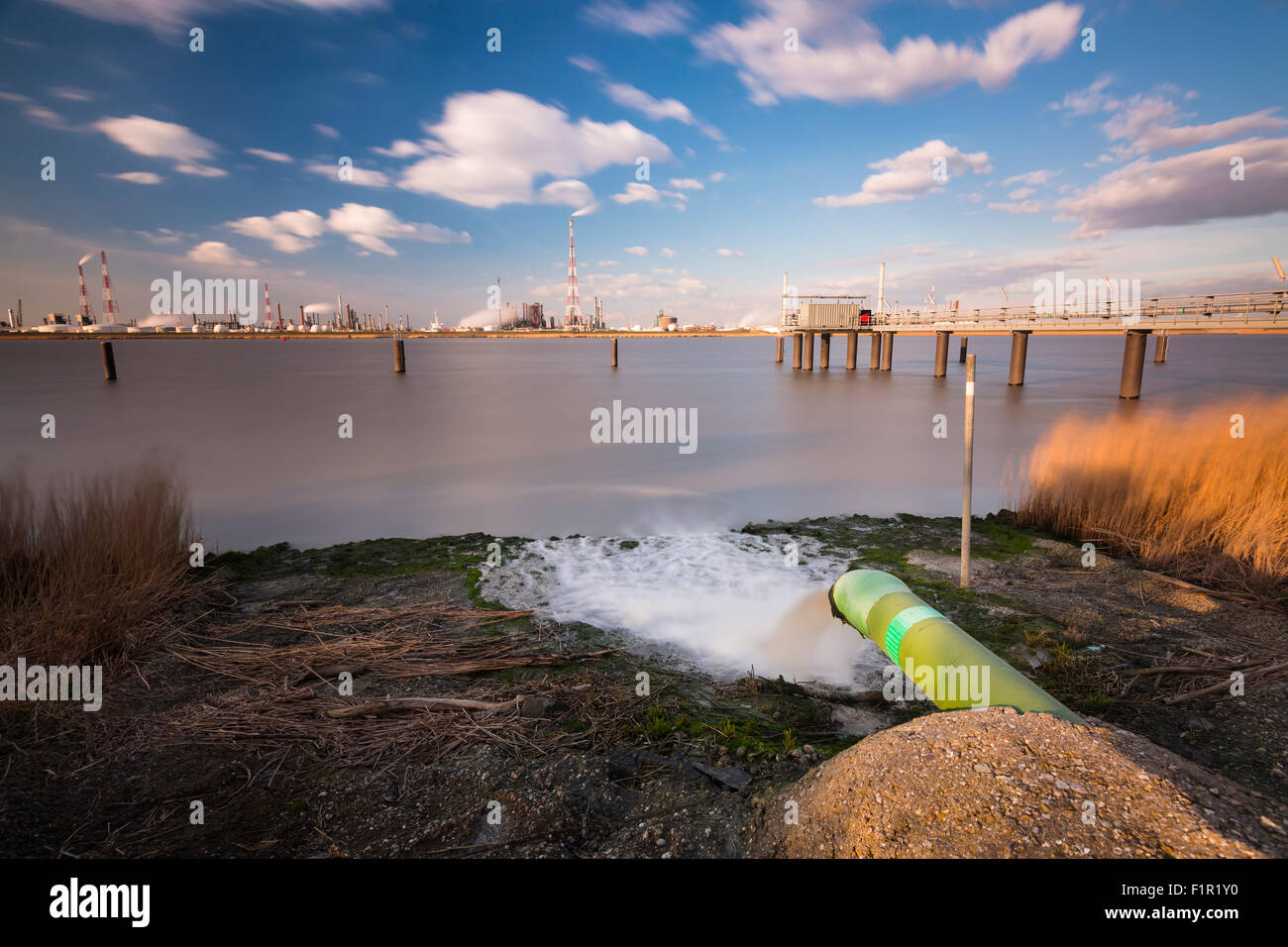 Long exposure shot of a wastewater pipe and a large oil refinery in the harbor of Antwerp, Belgium with blue sky and warm evenin Stock Photo