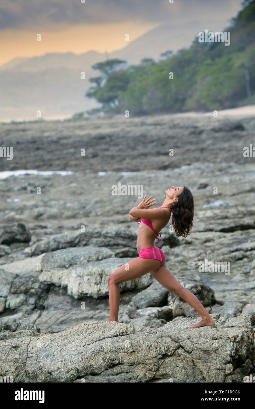 A young woman practising yoga on a wild, Pacific coastal rocky shore, surrounded by rainforest and unspoilt nature, Santa Teresa, Costa Rica Stock Photo