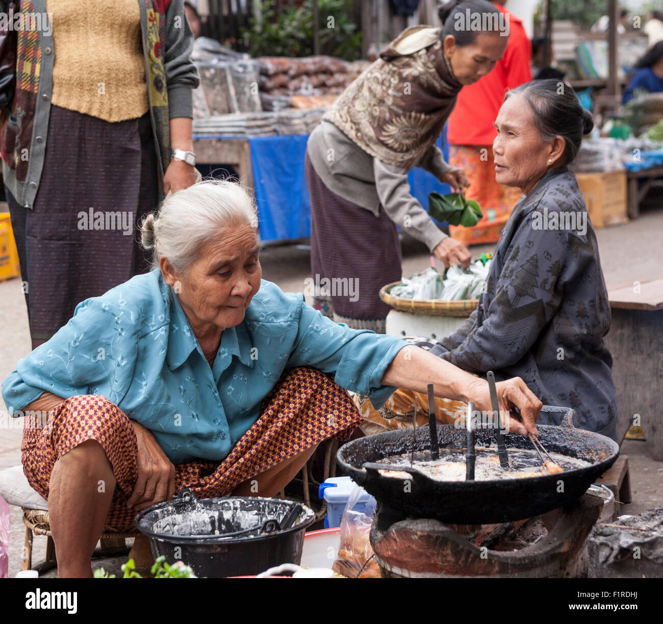 Elderly woman squats at a charcoal-fired wok to fry dough balls for sale in the street food market in Luang Prabang, Laos Stock Photo