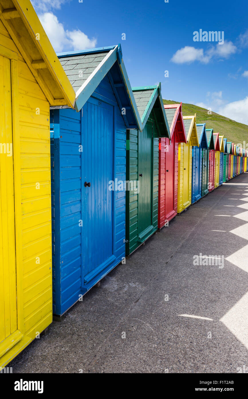 Row of colourful beach huts and their shadows with green hill backdrop, West Cliff Beach, Whitby, North Yorkshire, England, UK Stock Photo