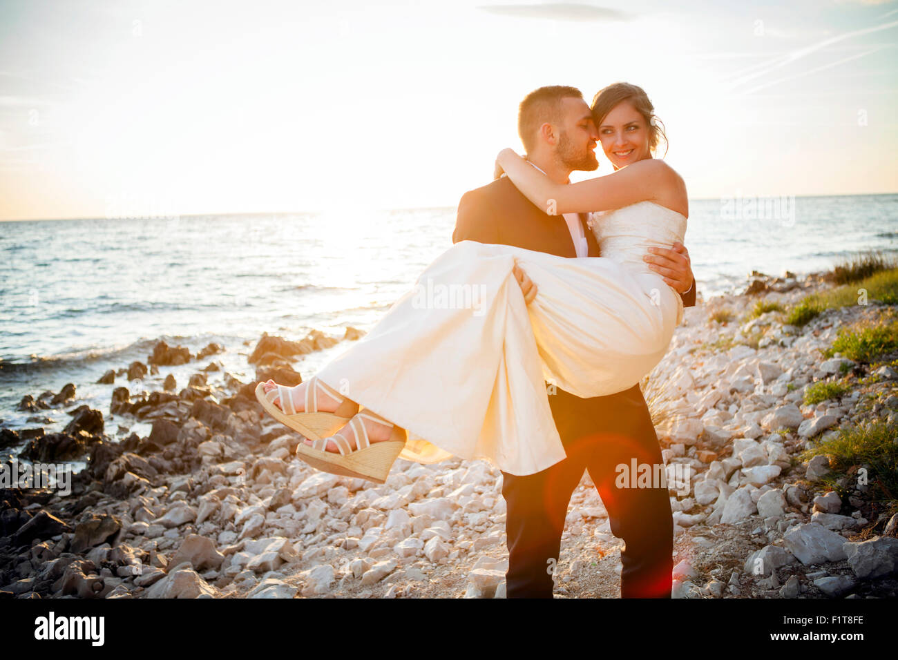 Groom carrying bride on pebble beach at sunset Stock Photo Alamy