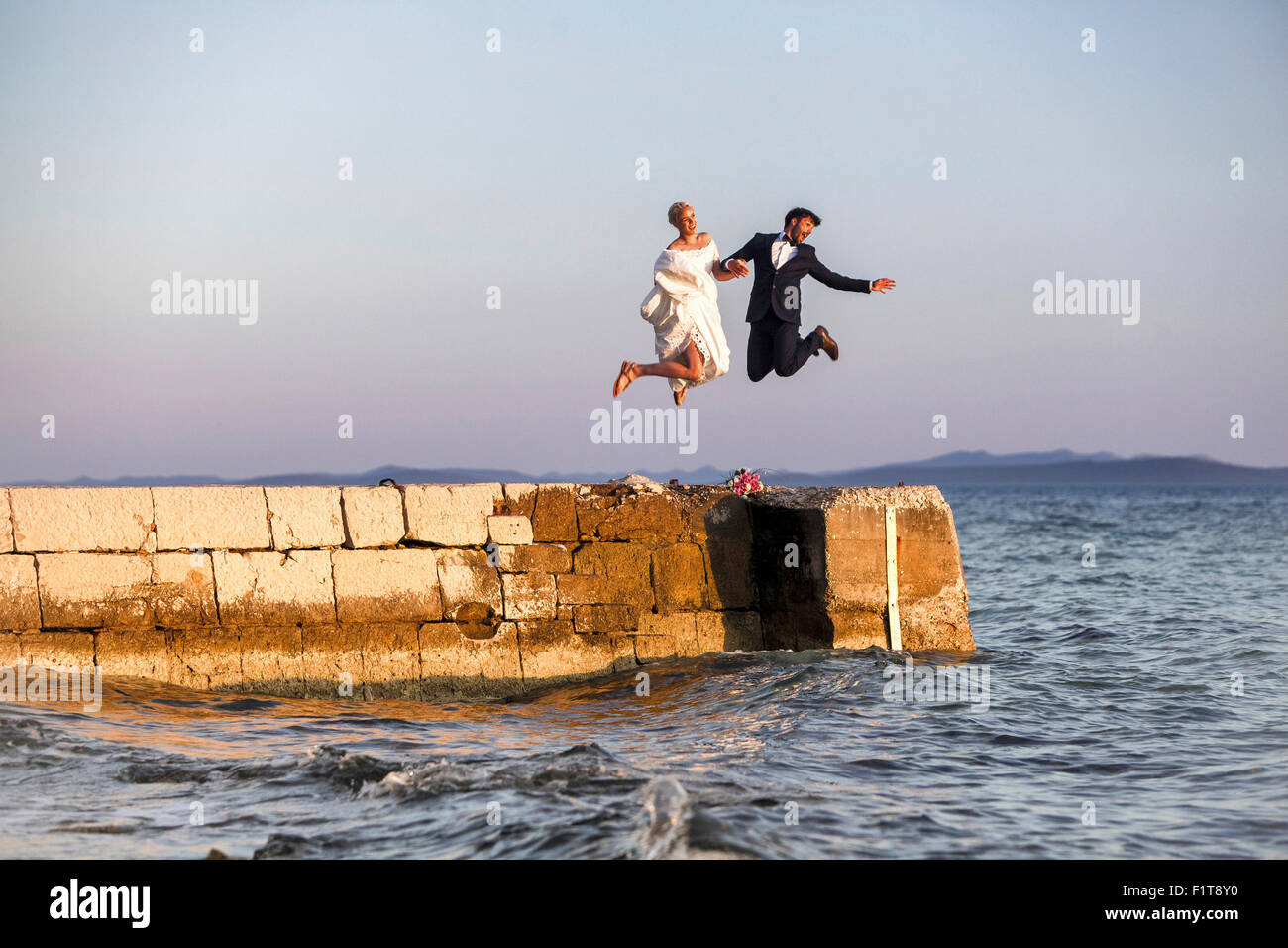 Bride and groom on pier jumping into the air Stock Photo