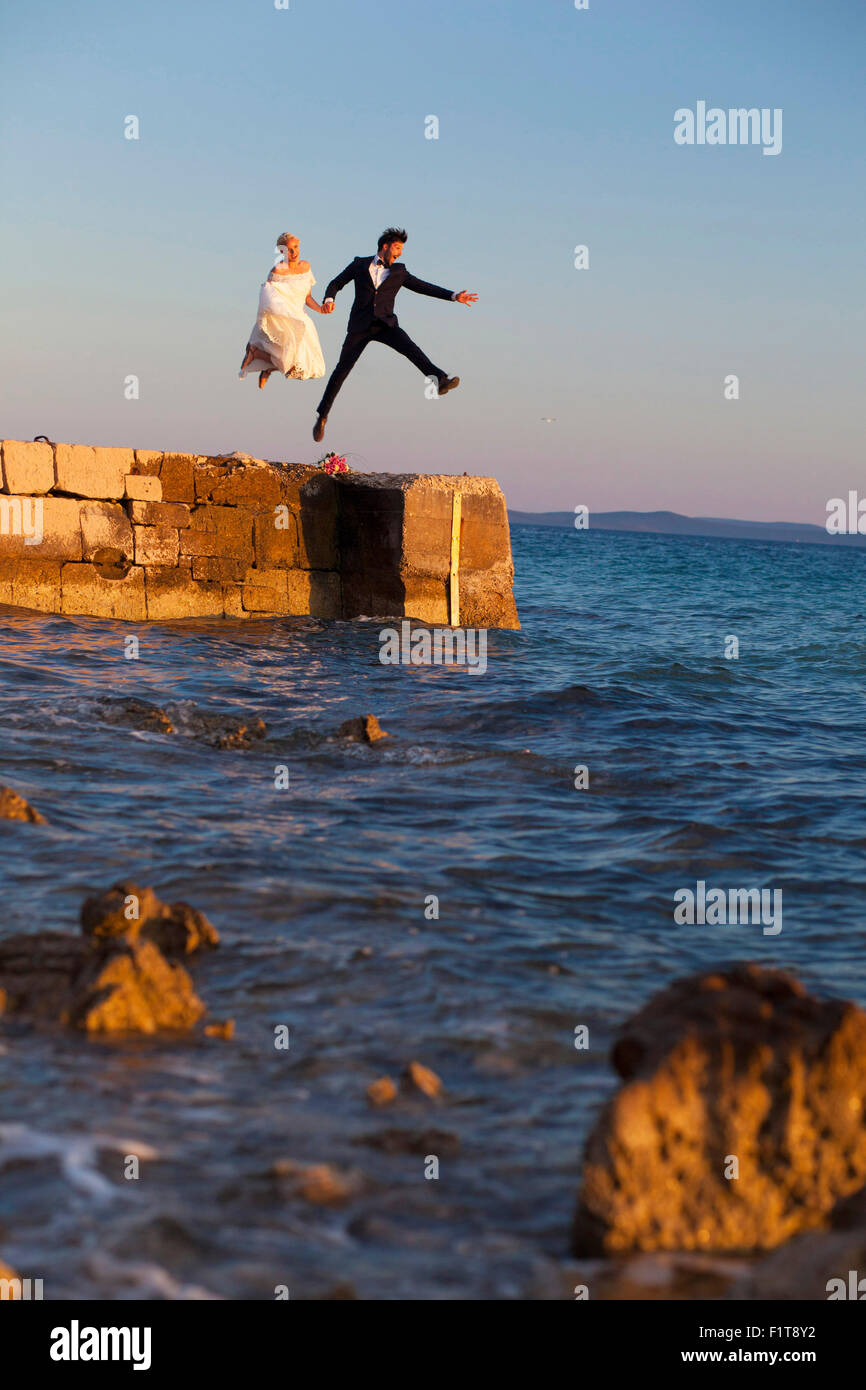 Bride and groom on pier jumping into the air Stock Photo