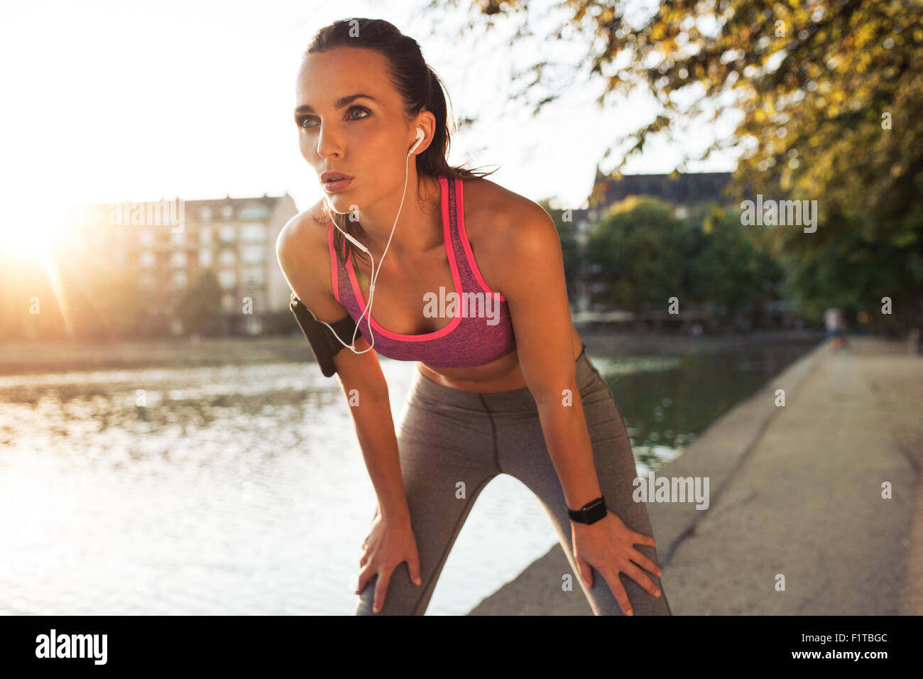 Female runner standing bent over and catching her breath after a running session along lake in city. Young sports woman taking b Stock Photo