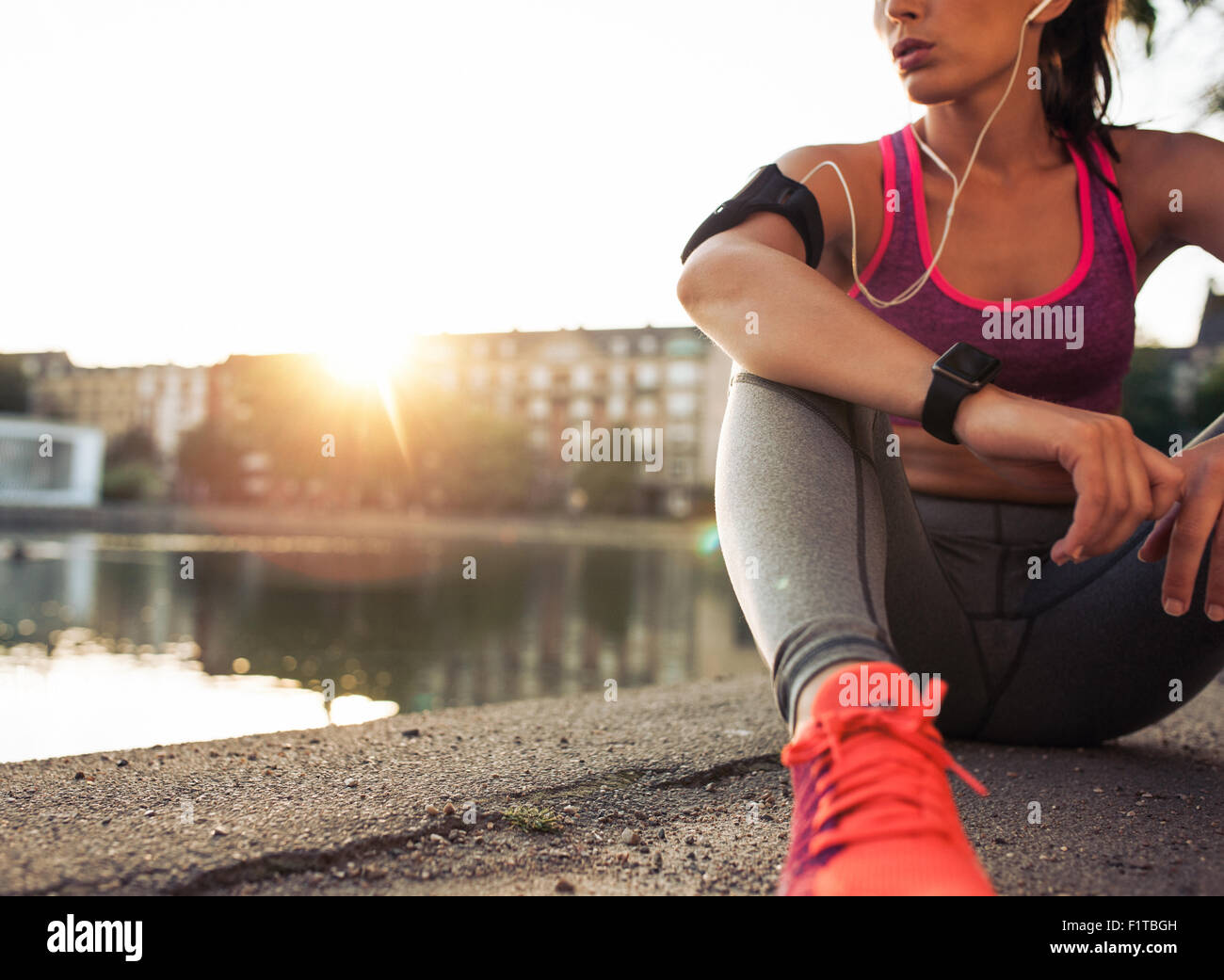 Young woman runner resting after workout session on sunny morning. Female fitness model sitting on street along pond in city. Fe Stock Photo