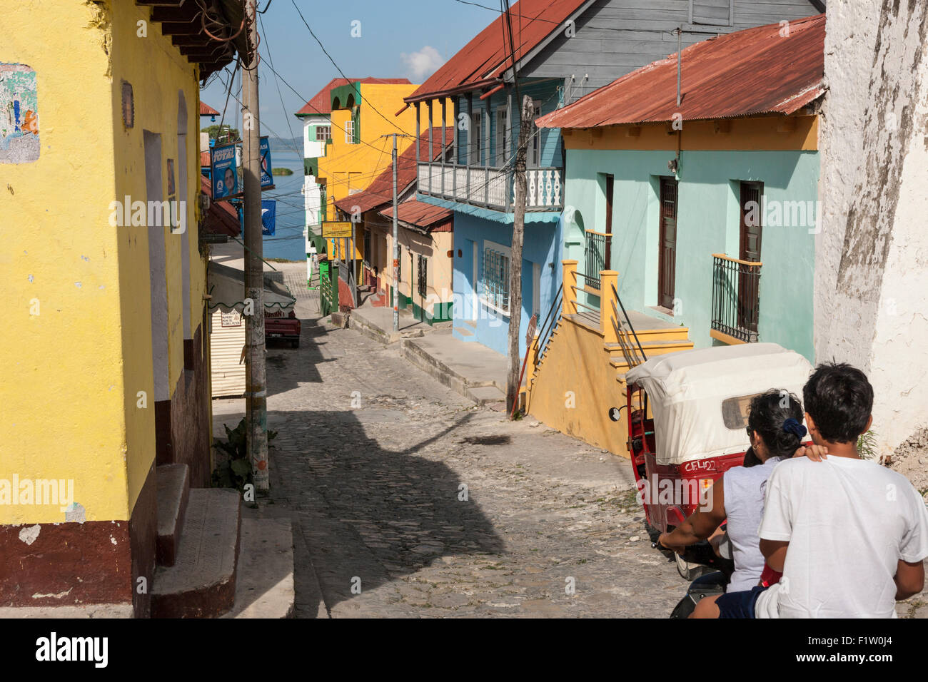 Motorcycle and jeep proceed down Calle de 10 Novembre past colorful houses in Flores, Guatemala. Lake Peten Itza visible beyond Stock Photo