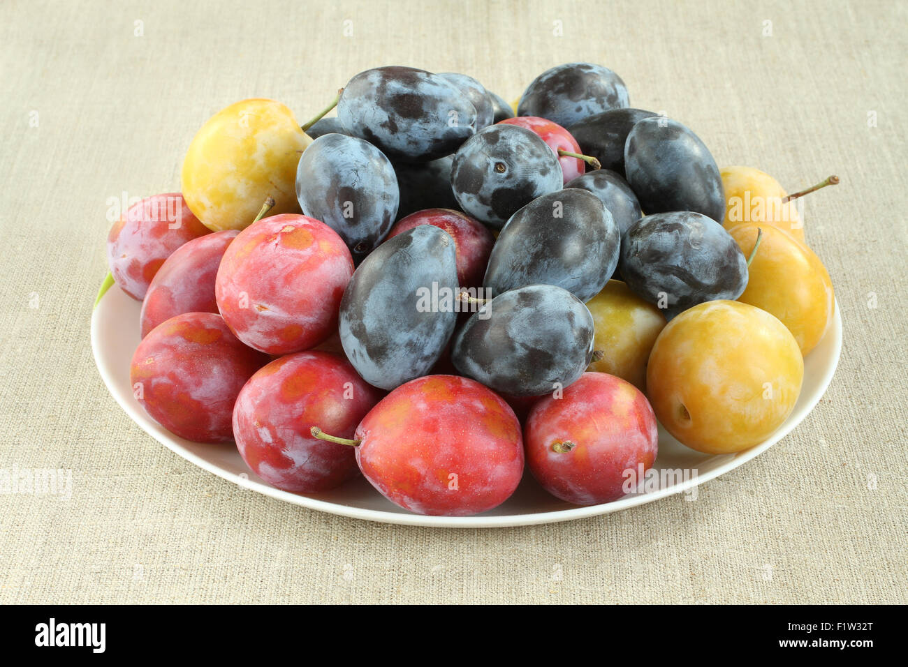 Ripe yellow, blue and pink plums in white plate on linen cloth. Closeup Stock Photo