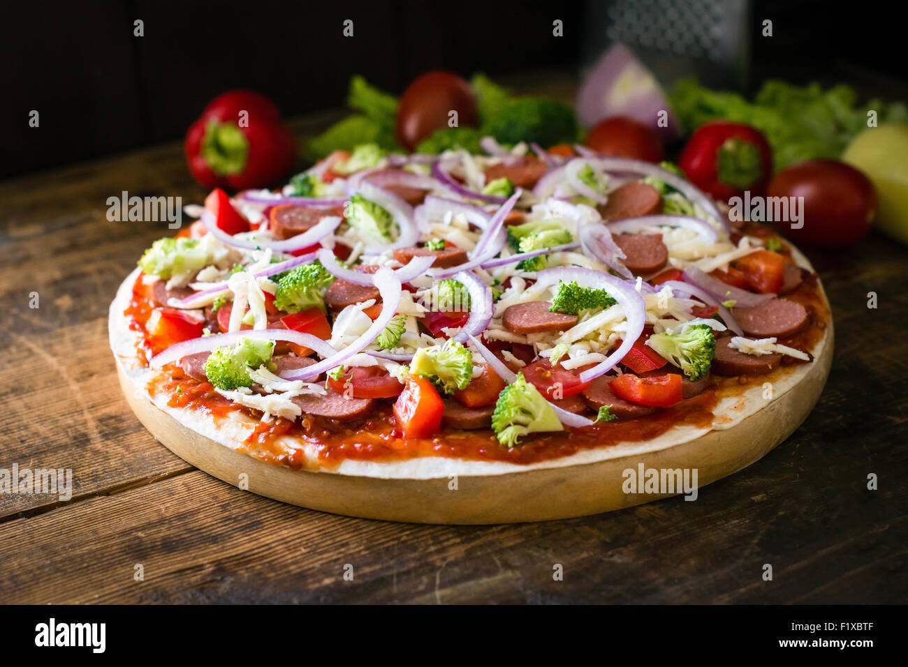 Thin crust pizza with sausages, pepper, tomatoes, broccoli and cheese on round wooden board Stock Photo