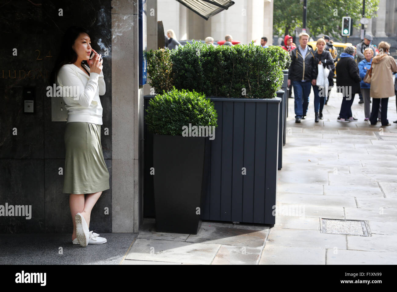 London 2015: An office worker takes a cigarette break outside her office building which fronts onto a busy city street. She using her mobile Stock Photo