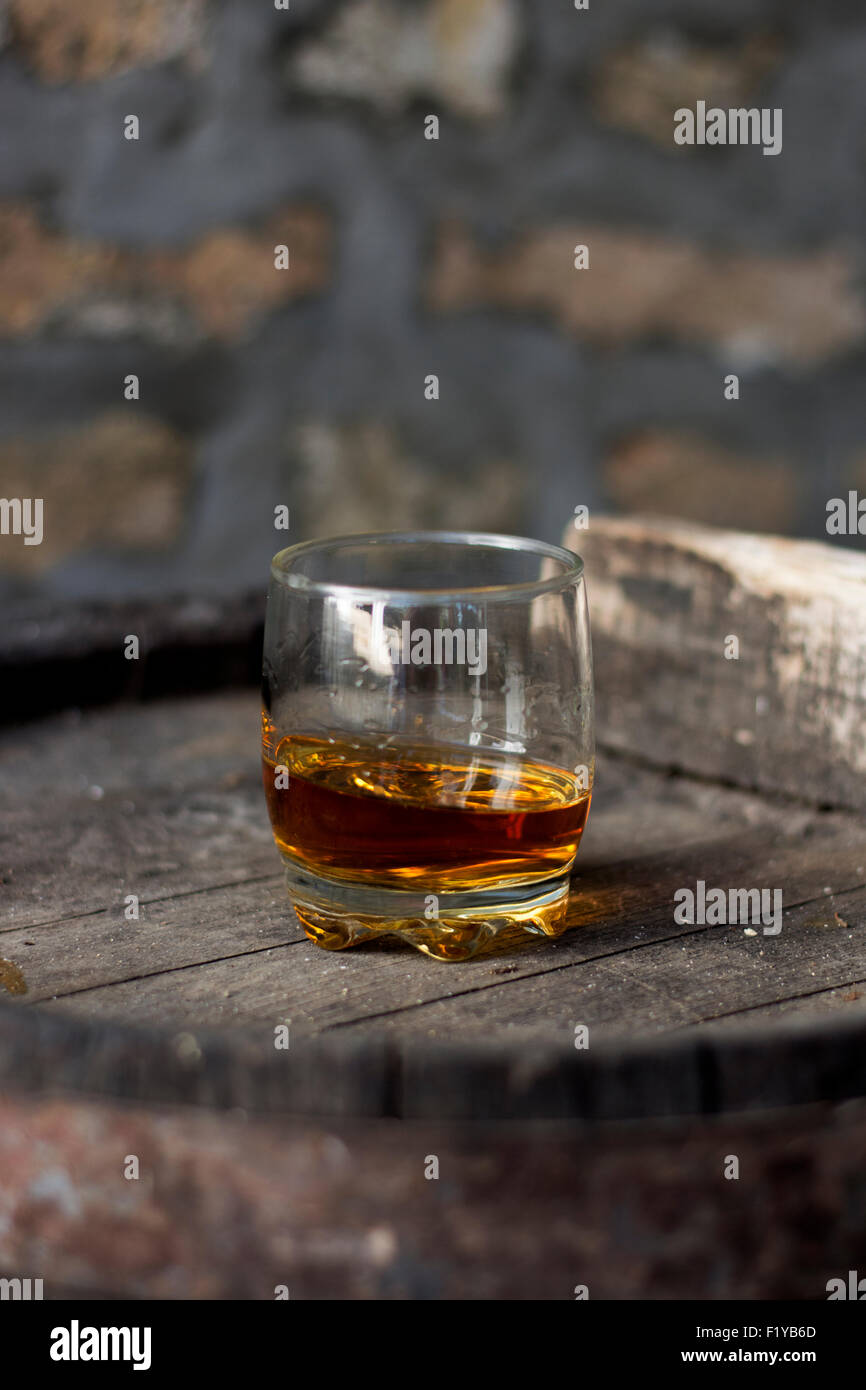Glass filled with whiskey, neat without ice, standing on a wooden barrel in distillery with barrels in the background. Stock Photo