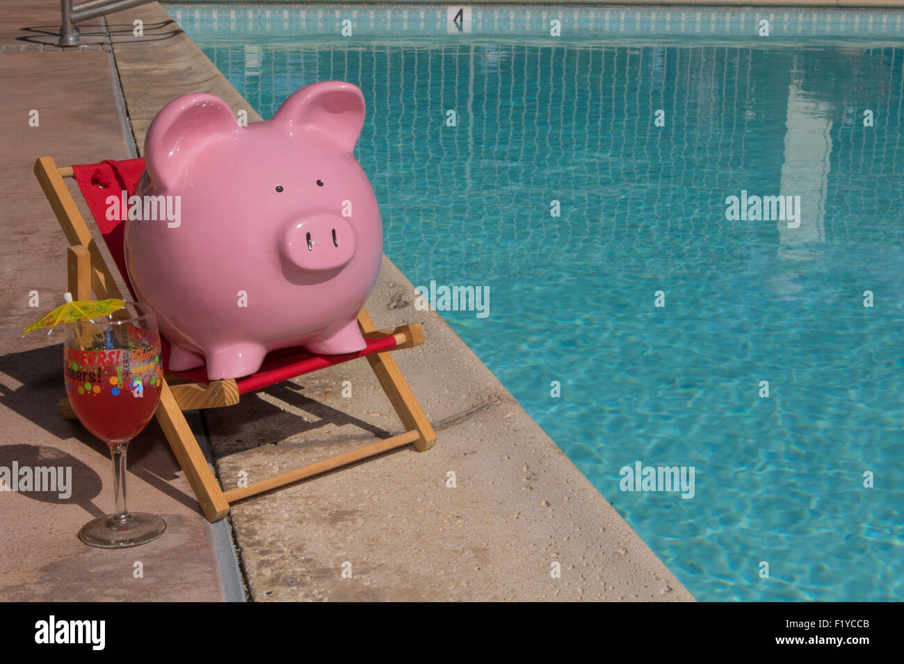 Pink piggy bank on a red deck chair relaxing by a pool on a hot summer day with a glass marked 'cheers'!  and cocktail umbrella Stock Photo